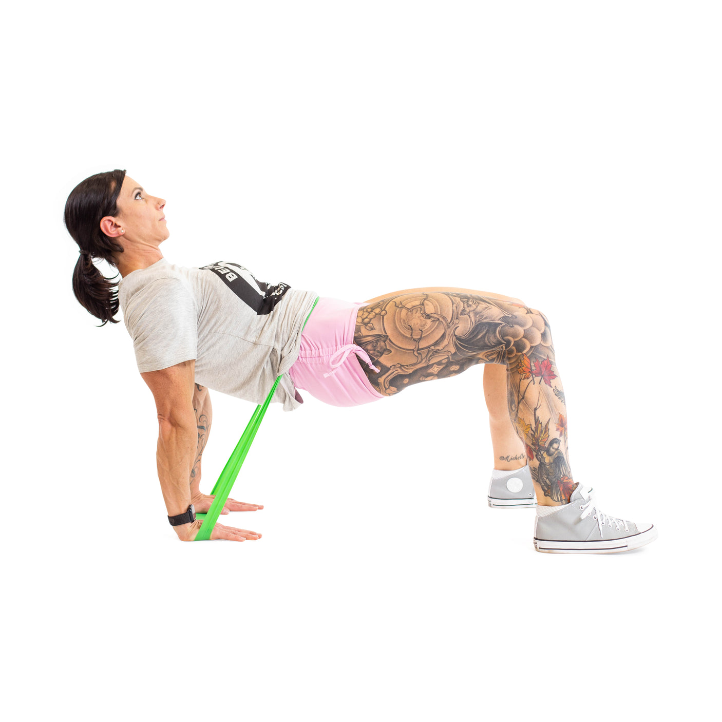 A woman with tattooed legs performs a glute bridge using Bells of Steel Mini Bands, dressed in pink shorts, a gray t-shirt, and gray sneakers against a white background.