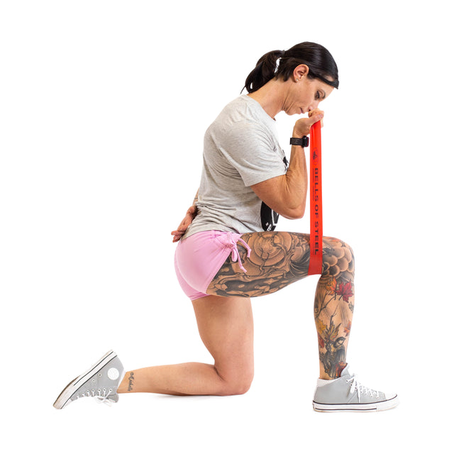 A tattooed person kneels on one knee, pressing a Bells of Steel Mini Band against their thigh while wearing a gray t-shirt, pink shorts, and gray sneakers—demonstrating gym accessories on a white background.