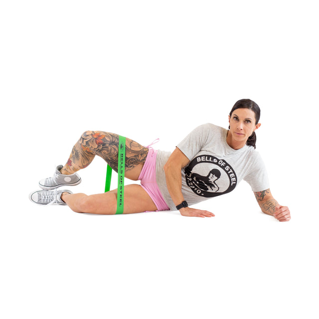 A person does clamshells with Bells of Steel Mini Bands (green) around their thighs, wearing a gray Bells of Steel t-shirt, pink shorts, and sneakers. Tattoos are visible on their arms and legs.