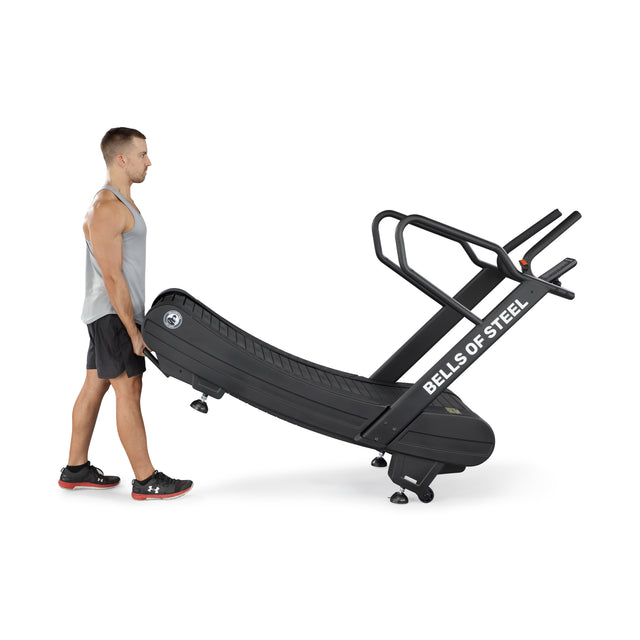 A man in athletic wear tilts and rolls the Blitz Magnetic Resistance Manual Treadmill by Bells of Steel using its rear wheels. The black, curved treadmill features handlebars and is angled upwards as he moves this sled push treadmill.