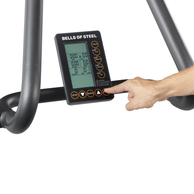 A hand presses a button on the digital control panel of the Blitz Magnetic Resistance Manual Treadmill by Bells of Steel, displaying workout stats. The panel is attached to a curved treadmill with black metal bars.