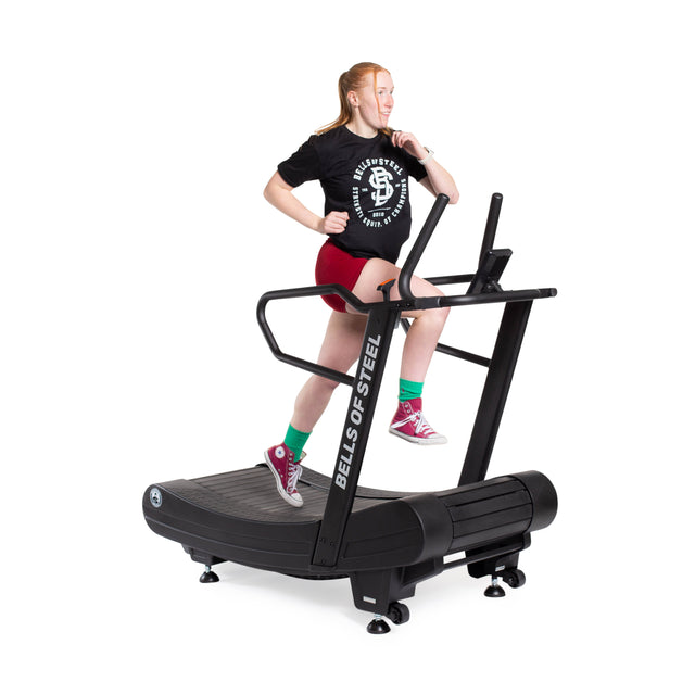 A woman, focused and energetic in a black T-shirt, red shorts, and colorful socks, runs on the Bells of Steel Blitz Magnetic Resistance Manual Treadmill—a non-motorized machine powering her workout.