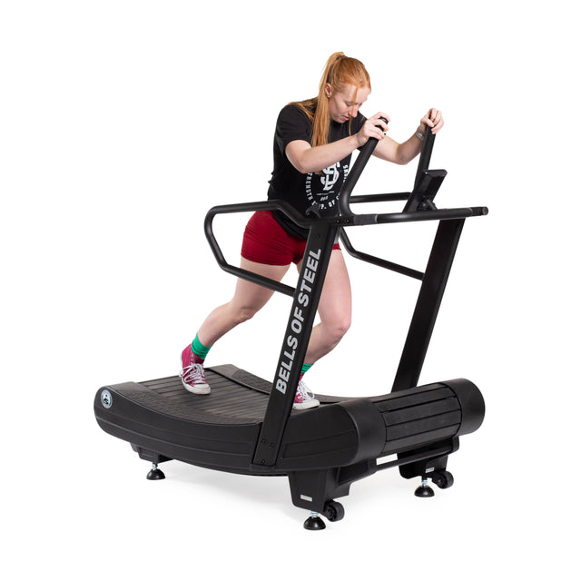 A person with long red hair, in a black shirt, red shorts, green socks, and red sneakers runs on the Blitz Magnetic Resistance Manual Treadmill by Bells of Steel, set against a white background.