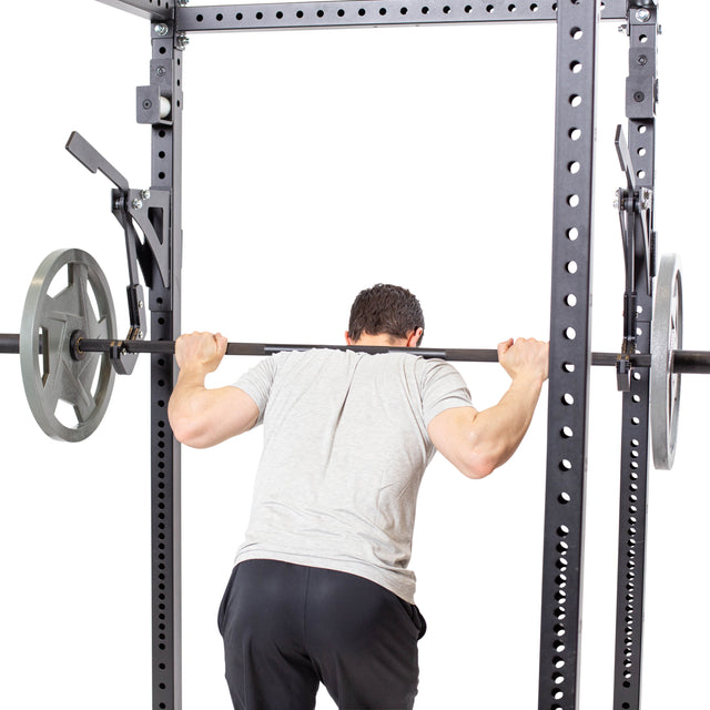 A man in a gray t-shirt and black pants performs a barbell squat using the Bells of Steel Monolift Rack Attachment, holding a weighted barbell across his upper back. Photo taken from behind against a white background.