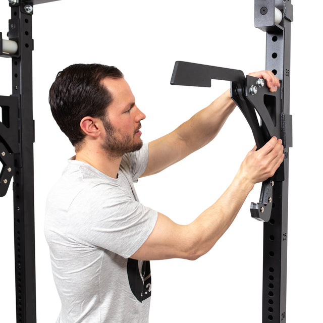 A man in a light gray t-shirt installs the black Bells of Steel Monolift Rack Attachment onto a tall, numbered gym rack. The space-saving setup stands out against the white background.
