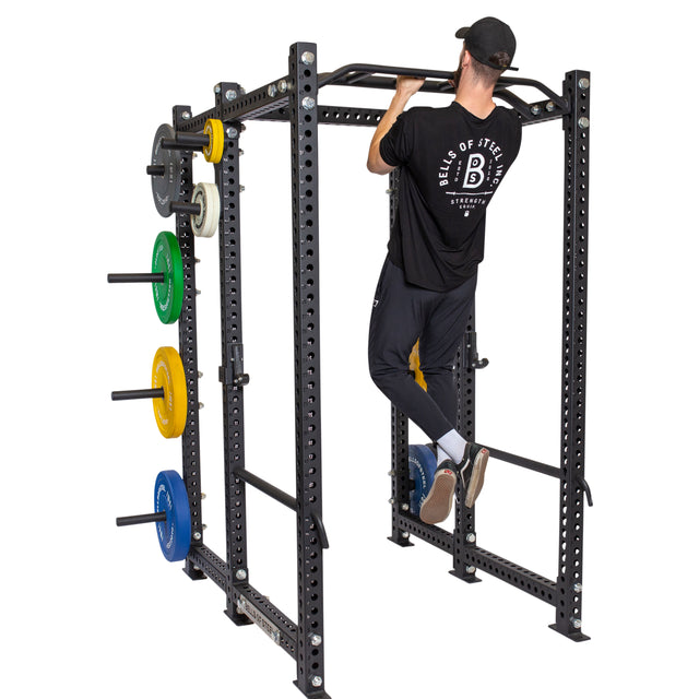 A man in a black shirt and cap does pull-ups on the Bells of Steel Multi-Grip Pull Up Bar attached to a power rack with colorful weight plates stored on its sides, showcasing his strength training routine.