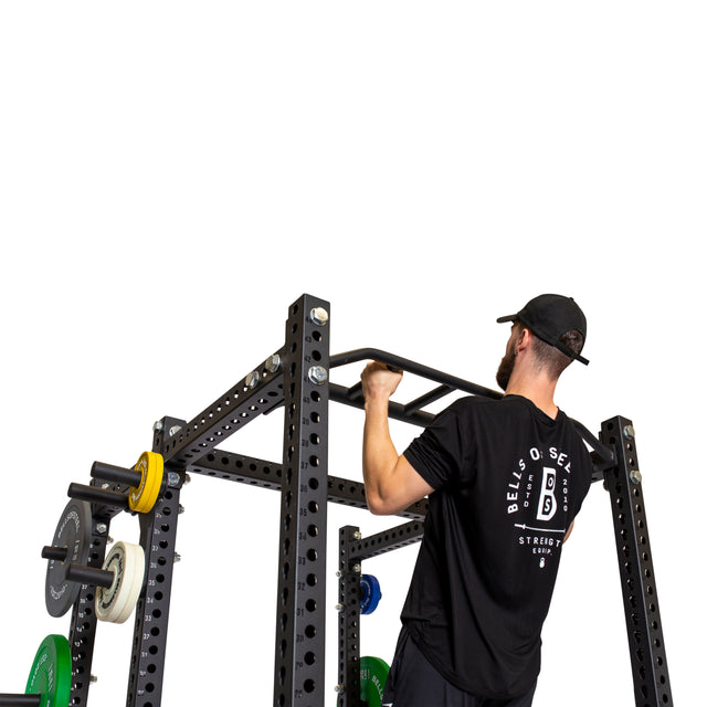 A man in a black cap and t-shirt performs a pull-up on the Bells of Steel Multi-Grip Pull Up Bar attached to a power rack with weight plates, against a white background—ideal for strength training.