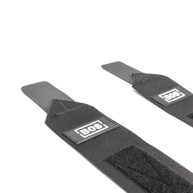 Close-up of two black Mighty Wrist Wraps by Bells of Steel, each with Velcro, a flat stiff tab, and rectangular brand labels. Perfect for wrist support during heavy lifts, they're laid out on a white surface.