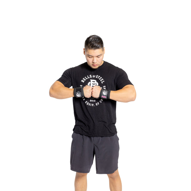 A man in a black T-shirt and shorts stands facing forward, looking down at his clasped hands. He wears Bells of Steel Mighty Wrist Wraps—essential for powerlifting support—on both wrists against a white background.