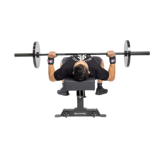 Wearing Bells of Steel Mighty Wrist Wraps for support, a person grips a barbell with both hands and performs a bench press on a weight bench labeled "BELLS OF STEEL" at its base.