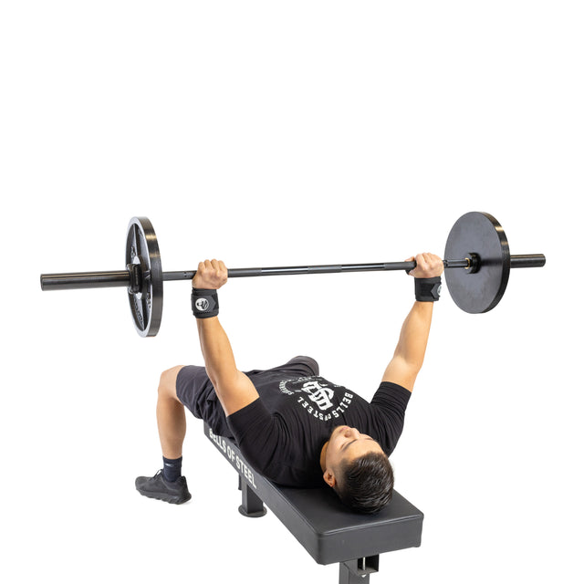 A person lies on a bench, gripping a barbell above their chest and preparing to bench press. They wear black shorts, a black shirt, and Bells of Steel Mighty Wrist Wraps for wrist support.