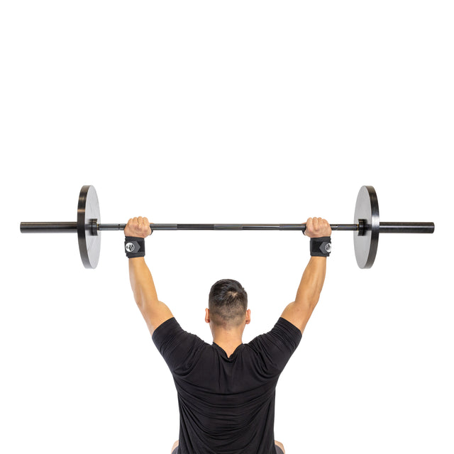 A man in a black shirt, wearing Bells of Steel Mighty Wrist Wraps, lifts a barbell overhead, seen from behind on a white background.
