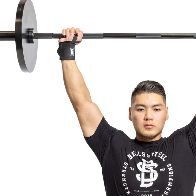 A man in a black Bells of Steel shirt lifts a barbell overhead with one arm, showcasing powerlifting essentials like the Mighty Wrist Wraps by Bells of Steel, against a white background.