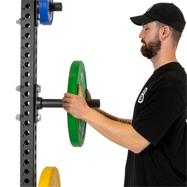 A man in a black t-shirt and hat loads a green Olympic plate onto the bar of a black power rack, with extra plates stored on Bells of Steel Bolt-On Plate Pegs mounted below.