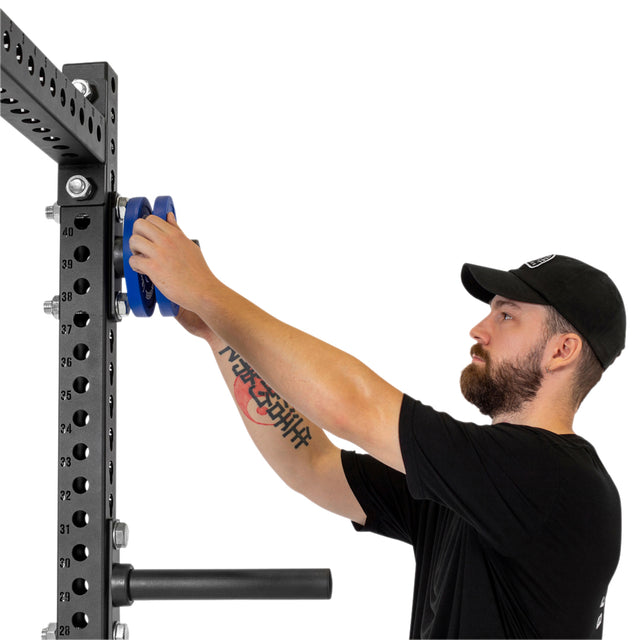 A man in a black cap and shirt loads blue plates onto Bells of Steel Bolt-On Plate Pegs, which are mounted on the side of a metal power rack with numbered holes against a white background.