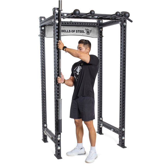 A person in a black t-shirt and shorts adjusts a barbell on a Bells of Steel power rack with multiple adjustment holes, featuring the Vertical Mount Barbell Holder Rack Attachment for efficient barbell storage inside the rack.