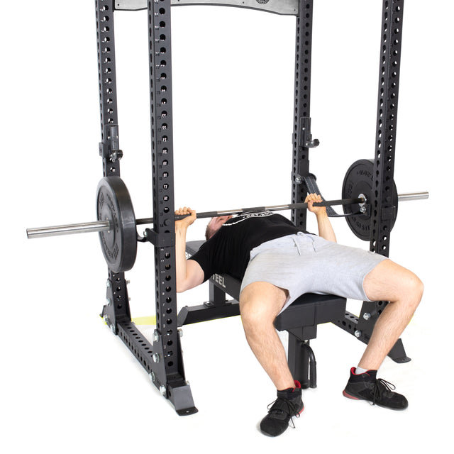 A man prepares to bench press under a barbell in the Bells of Steel Manticore Flat Foot Power Rack - Prebuilt (3" x 3", 1" Holes), wearing a black tee, gray shorts, and black shoes, shown on a white background—perfect for home gyms.