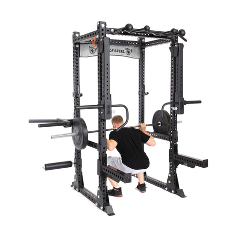 A man squats with a barbell inside the Bells of Steel Manticore Flat Foot Power Rack - Prebuilt (3" x 3", 1" Holes), equipped with attachments and weight plates, perfect for home gyms, set against a white background.