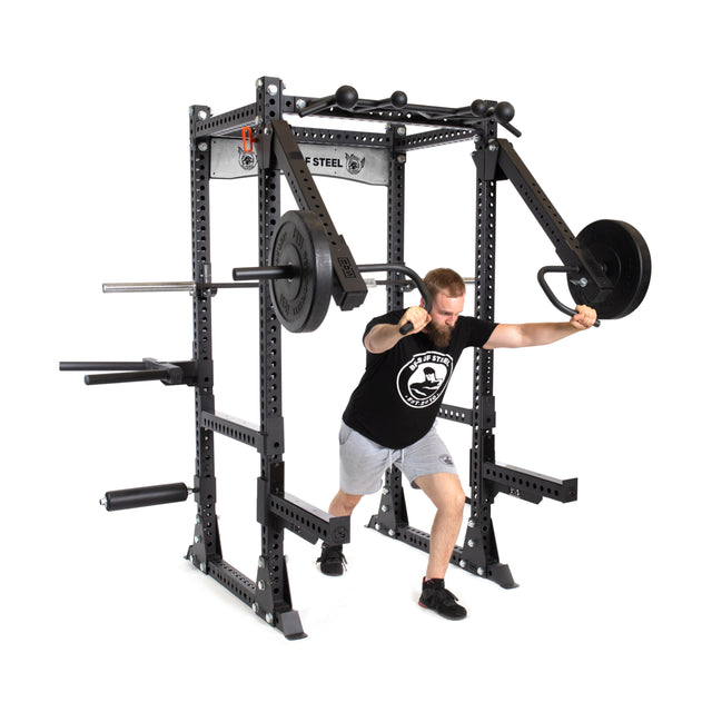 A man in a black t-shirt and gray shorts uses the Manticore Flat Foot Power Rack - Prebuilt (3" x 3", 1" Holes) by Bells of Steel to perform a push exercise, lunging forward while pushing the handles and loading weight plates.