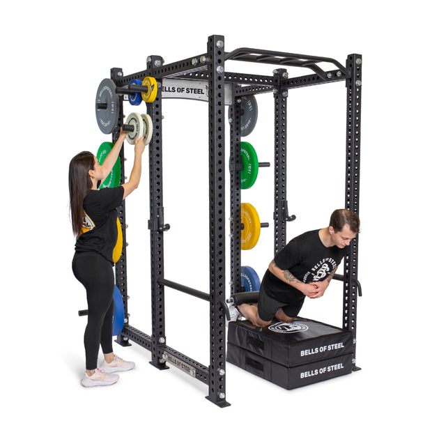 A woman adjusts weight plates on a Bells of Steel Manticore Six Post Power Rack Builder with 3"x3" uprights, as a man exercises on a pad nearby. The modular rack features colorful weights. Both wear black athletic attire.