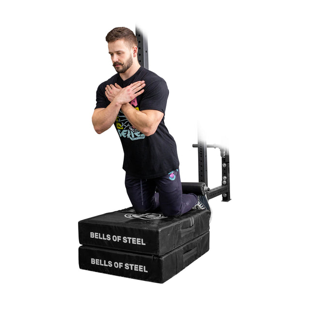 A man kneels on the Bells of Steel Hydra Nordic Curl Package, arms crossed over his chest, performing Nordic hamstring curls with support from its split squat attachment and rack.