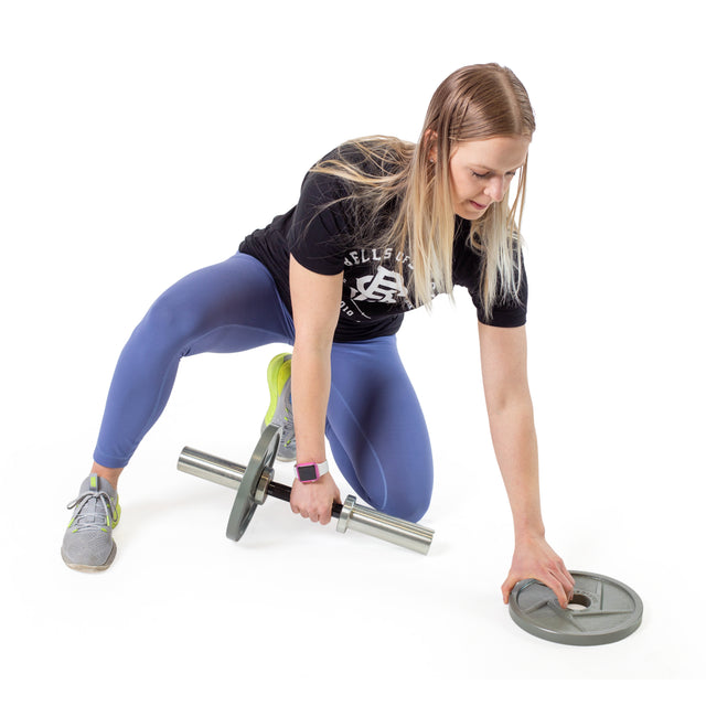A woman in a black T-shirt and blue leggings kneels on one knee, using the Bells of Steel Loadable Dumbbell Handle and reaching for a weight plate—ideal for any home gym setup against a white background.
