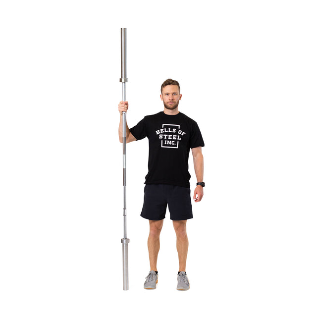 A man in a black Bells of Steel t-shirt and shorts stands on a white background, holding a Bells of Steel Olympic Weightlifting Barbell vertically with one hand.