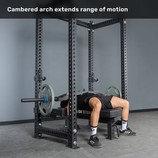 A person uses the Bells of Steel Arch Nemesis Swiss Bar in a power rack, performing a bench press with a cambered arch for greater range of motion. Specialty barbells like this are excellent for boosting your bench. Text: "Cambered arch extends range of motion.
