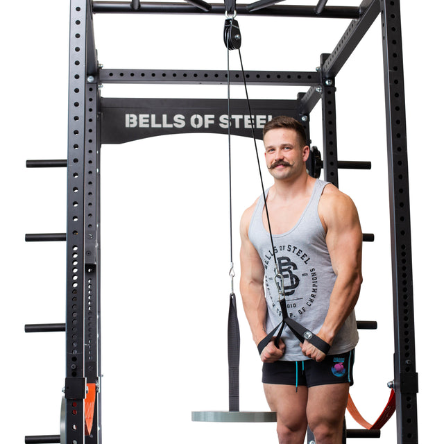 A muscular, mustached man in a gray tank top and black shorts stands in a Bells of Steel power rack, gripping the Cable Pulley attachment and preparing for tricep pushdowns with the portable machine.
