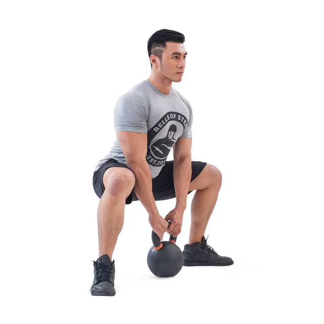 A man in a grey t-shirt and black shorts does a sumo squat while holding a Bells of Steel Powder Coated Kettlebell with both hands against a white background.
