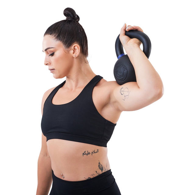 A woman in a black sports bra and leggings holds a Bells of Steel Powder Coated Kettlebell over her shoulder, showing its strong grip design against a white background. Her dark hair is in a bun and she has a script tattoo on her side.