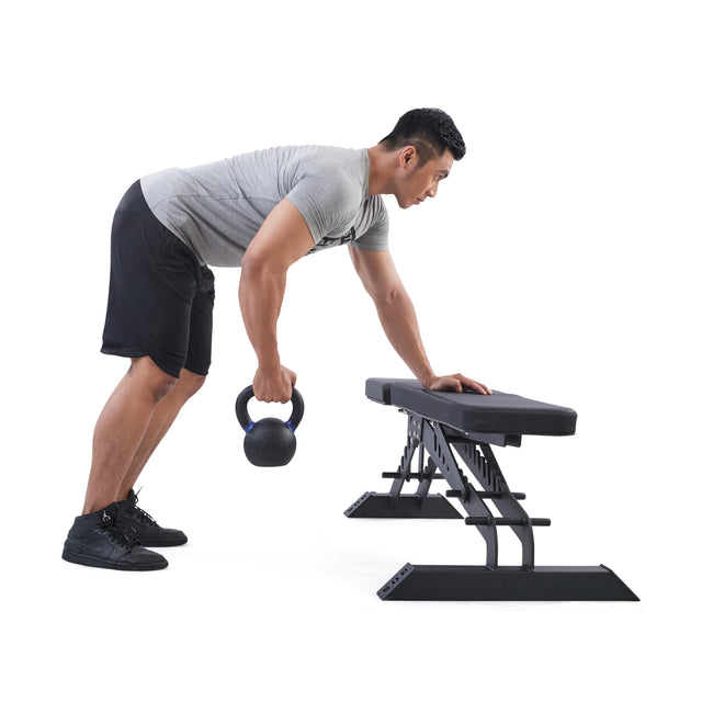 A man in a gray shirt and black shorts performs a bent-over row with a Bells of Steel Powder Coated Kettlebell, bracing himself on a bench for support and lifting with one hand for enhanced grip and rust resistance.