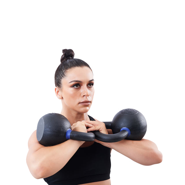 A woman with dark hair in a bun grips a Bells of Steel Powder Coated Kettlebell close to her chest, wearing a black sports bra against a plain white background.