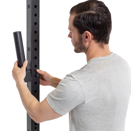 A man in a light gray t-shirt installs Bells of Steel Pin Plate Pegs onto a vertical steel rack with numbered holes, organizing his Olympic weight plate storage.