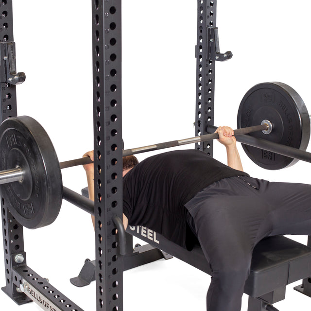 Wearing black, a person lies on a bench press inside a power rack, gripping a loaded barbell. Bells of Steel Pin Pipe Safeties are set for protection as they prepare to lift the weights.