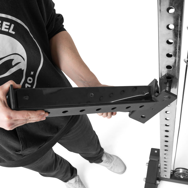 Wearing a black sweatshirt and white sneakers, someone adjusts a Bells of Steel Functional Trainer Cable Tower. They carefully align the metal bracket attachment with the upright post holes to enhance their workout setup.