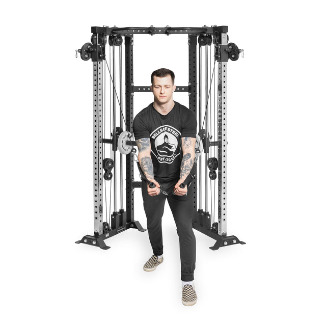 A tattooed individual in a black shirt and checkered shoes uses the Bells of Steel Functional Trainer Cable Tower for cable crossover exercises, maintaining focus amidst the array of weights.