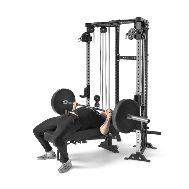 In their home gym, someone performs a bench press using a barbell on a flat weight bench next to the Bells of Steel Functional Trainer Cable Tower, which includes adjustable pulleys. They sport black athletic wear and checkerboard shoes for optimal style and function during the workout.