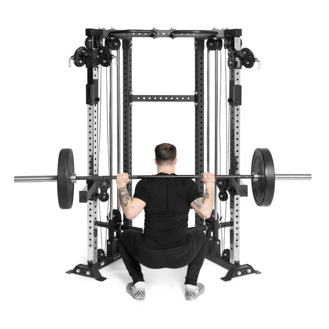 Clad in black, a person squats with a barbell supported by the Bells of Steel Functional Trainer Cable Tower, using its weight plates on each side.