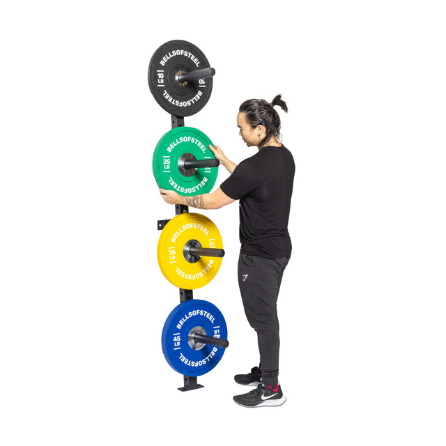 Wearing black athletic gear, a person arranges colorful weight plates on the Bells of Steel Wall-Mounted Plate Storage Rack, set against a white background—ideal for organized home gym storage.