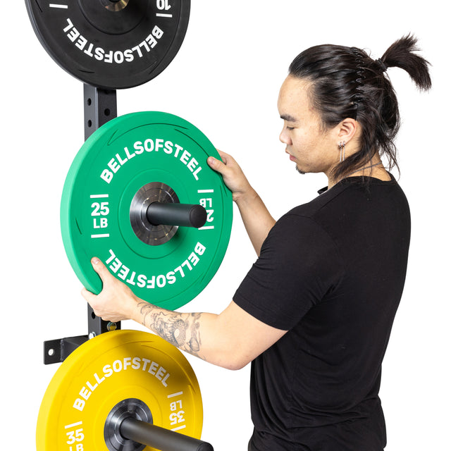 A person in a black shirt places a green 25 lb Bells of Steel plate onto the Bells of Steel Wall-Mounted Plate Storage Rack, which also organizes black and yellow weight plates in their home gym.