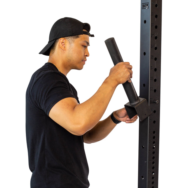 A man in a black shirt and backward cap uses a steel tool to adjust Bells of Steel Pin Plate Pegs on a tall black metal rack with numbered holes.