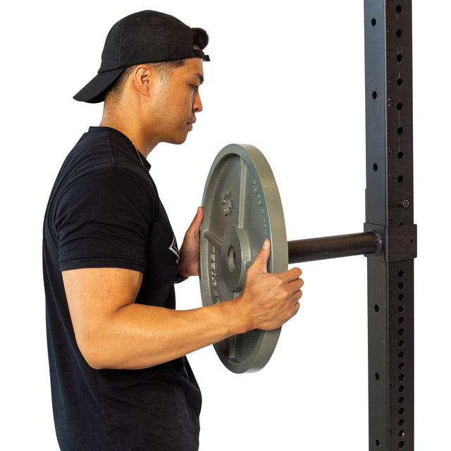 A man in a black shirt and backward cap loads a weight onto a barbell on a squat rack with Bells of Steel Pin Plate Pegs for Olympic plate storage. The background is white.