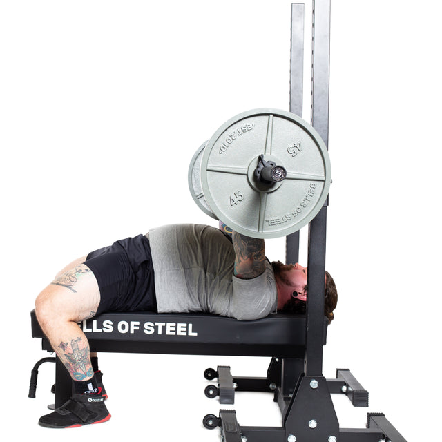 A tattooed person lies on a bench press, lifting a Bells of Steel Powerlifting Bar. The side view highlights their form during the exercise.