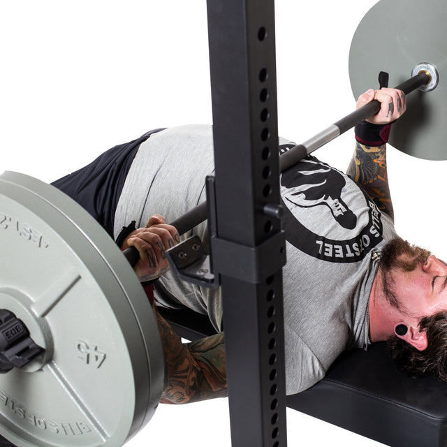 A tattooed man grips a Bells of Steel Powerlifting Bar, preparing to bench press on a gym bench. The bar is loaded, part of the rack is visible, and the background is white.