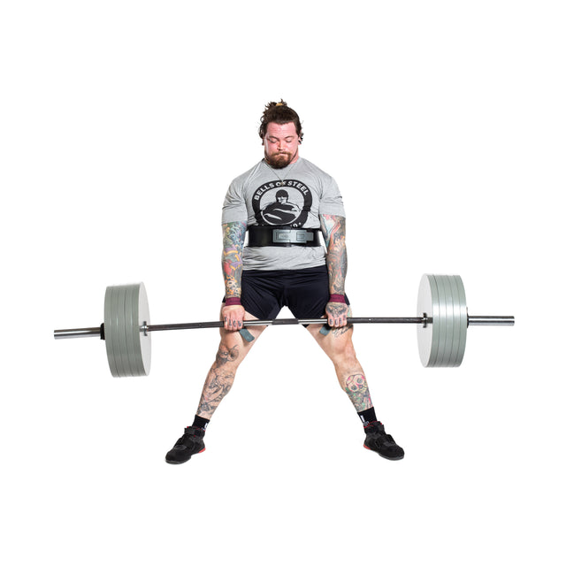 A tattooed, bearded man lifts the heavily loaded Bells of Steel Powerlifting Bar while wearing a weightlifting belt, gray t-shirt, and black shorts against a plain white background.