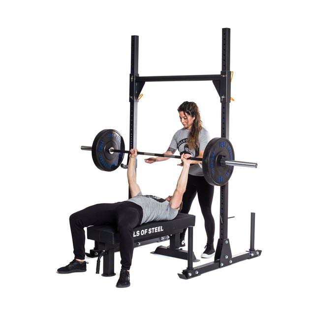 A person performs a barbell bench press on a weight bench with the Bells of Steel Powerlifting Bar, while another stands behind to spot. Both are dressed in athletic wear against a plain white background.