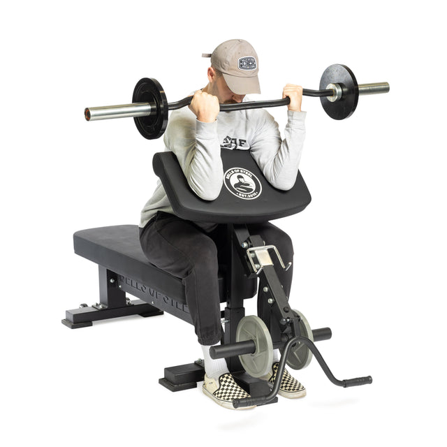 Wearing a cap, sweatshirt, and checkered shoes, a person performs a barbell curl on Bells of Steel Bench Attachments in a gym, using a black and silver weightlifting setup.