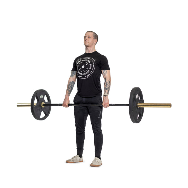 A person with arm tattoos stands on a white background, wearing a black t-shirt and pants, while lifting a barbell featuring ergonomic handles and Rubber Coated Iron Weight Plate Sets by Bells of Steel.