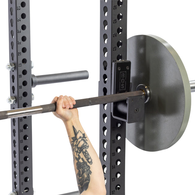 A tattooed arm grips a barbell loaded with weights on a Bells of Steel squat rack featuring numbered adjustment holes and Roller J-Cups with durable nylon rollers, set against a white background that emphasizes the hand, barbell, and rack.